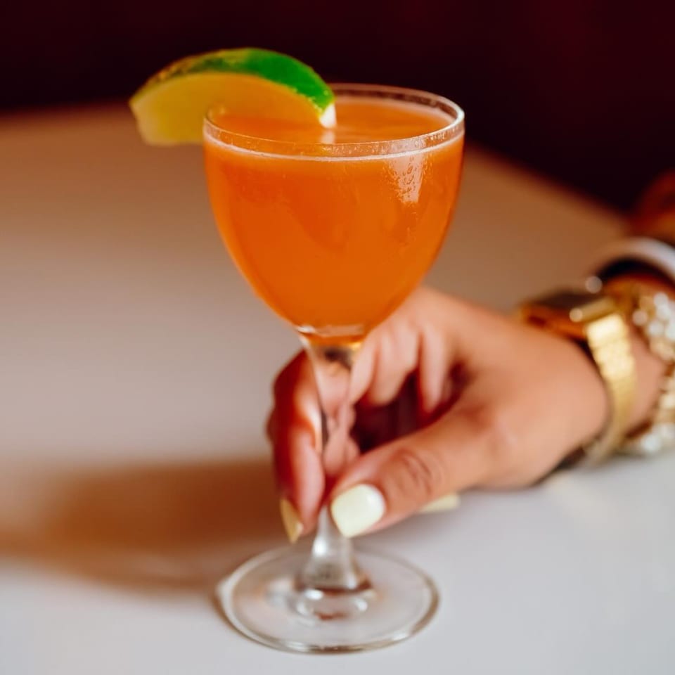 A hand with white nail polish holds a vibrant orange cocktail garnished with a lime wedge.