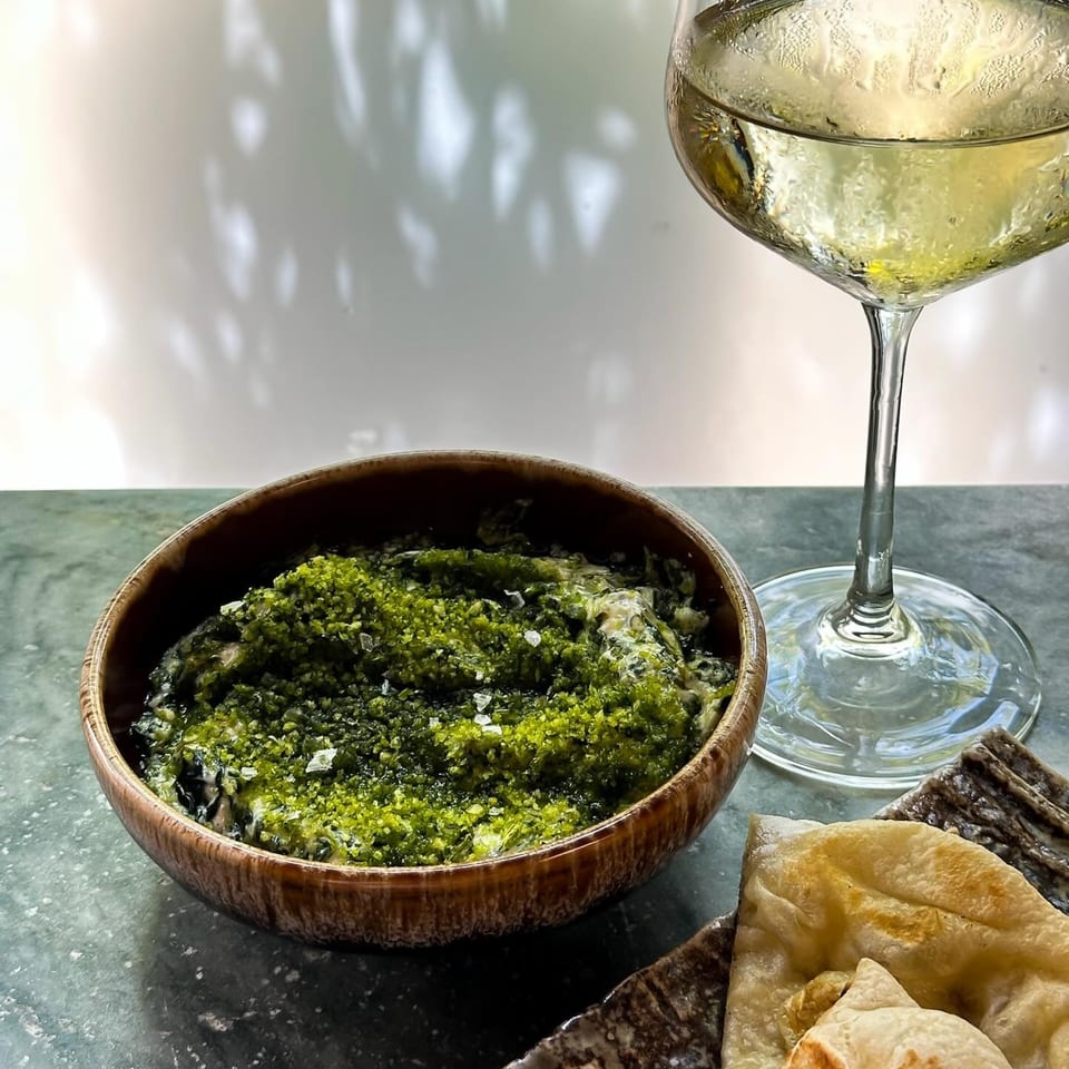 A bowl of green dip with flatbread and a glass of white wine on a speckled table.