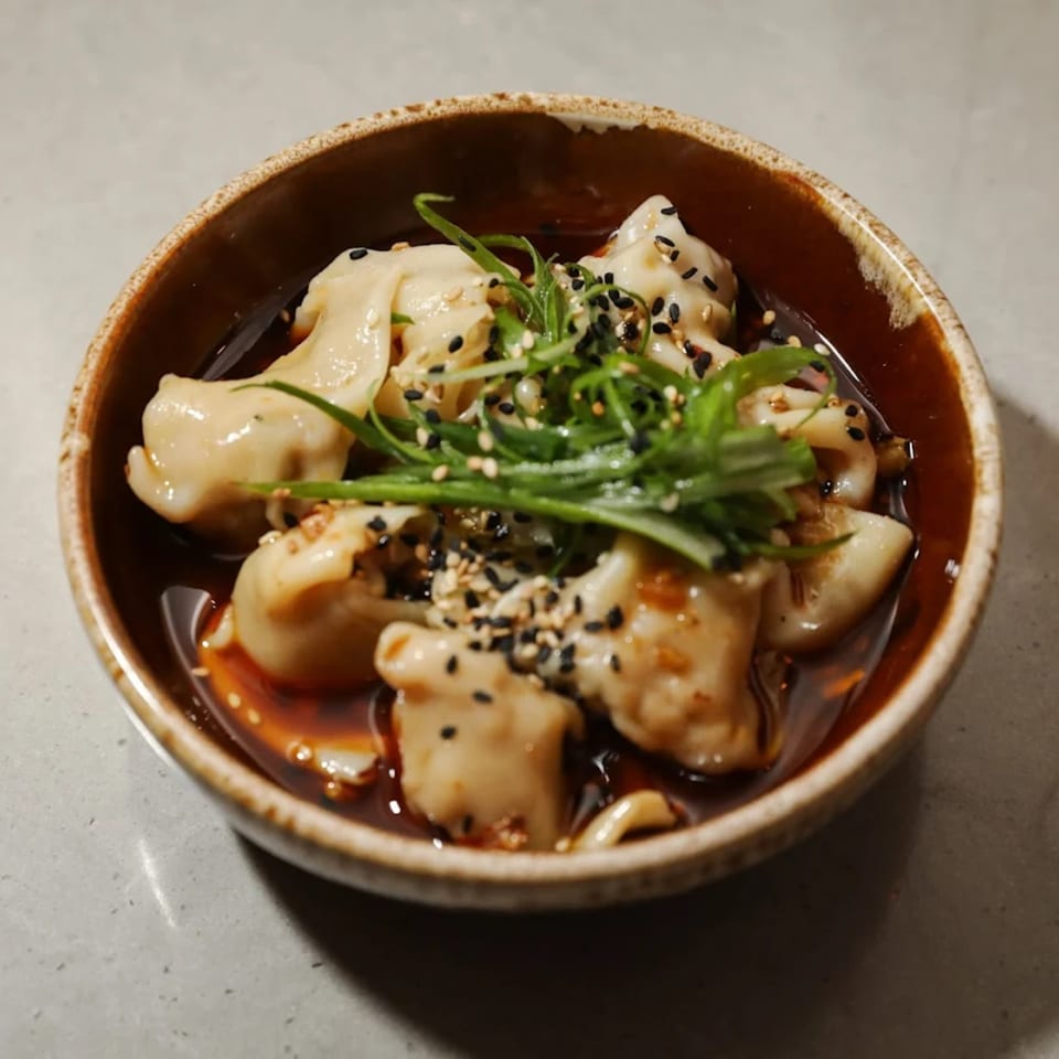 A bowl of Edgemar Restaurant + Lounge's dumplings in sauce, topped with green onions and sesame seeds.