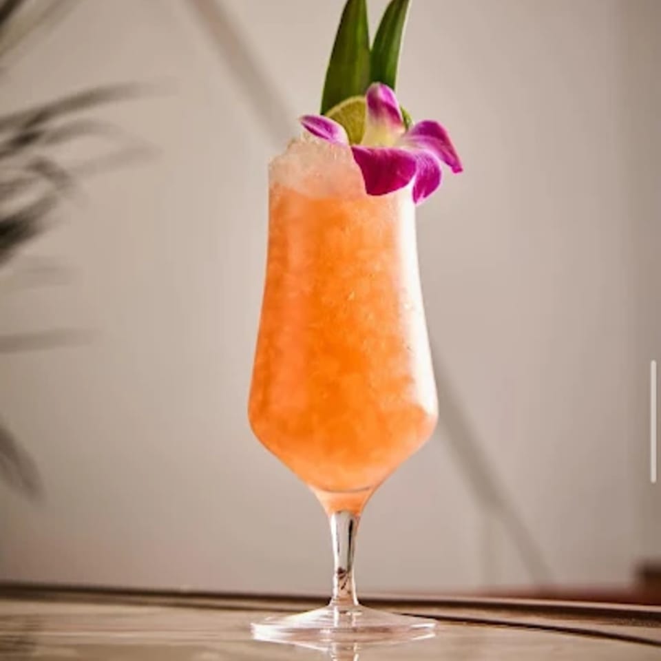 An orange cocktail with crushed ice, garnished with a lime wedge, green leaves, and a purple orchid.