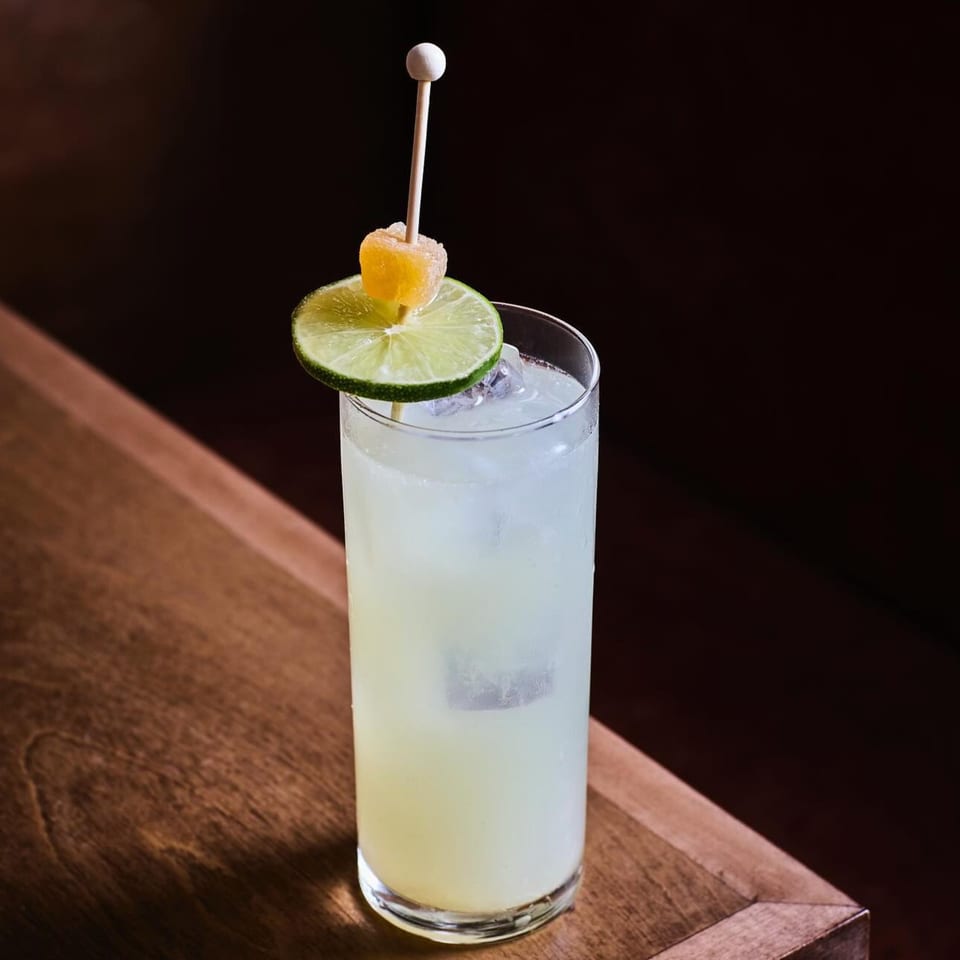 A tall glass of light yellow cocktail with ice, garnished with a lime slice and candied ginger.