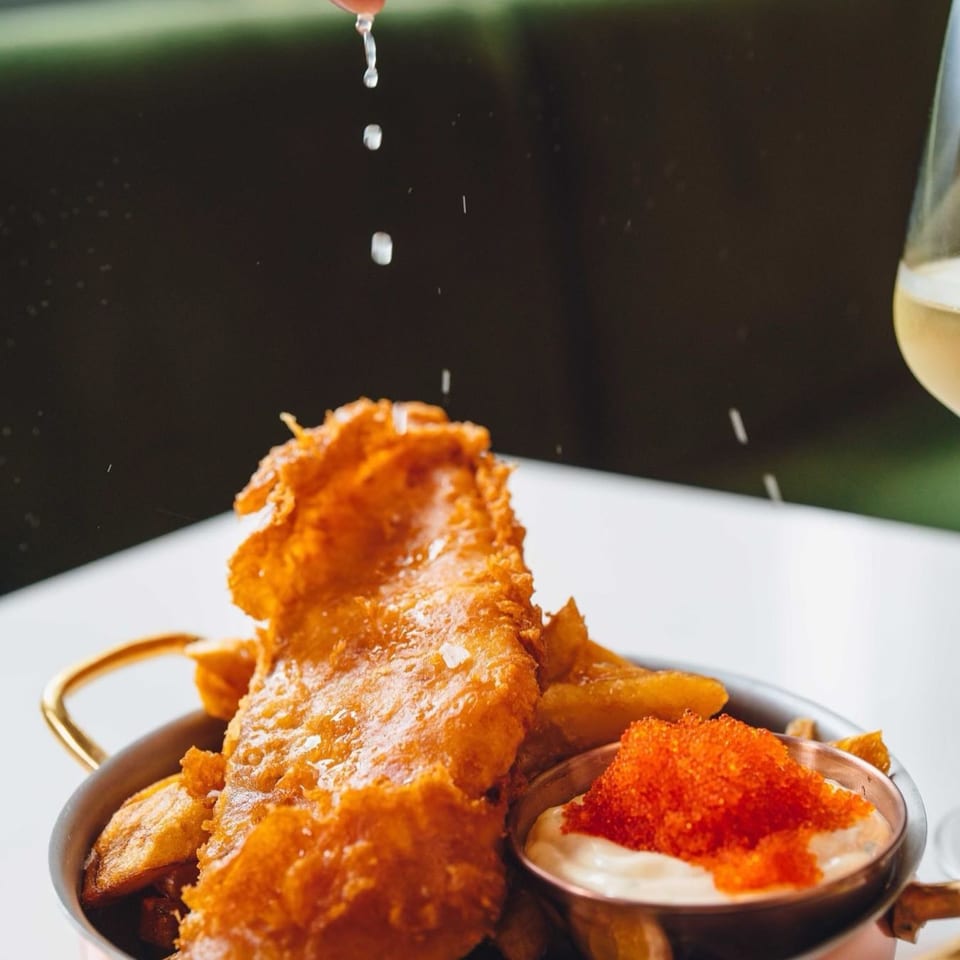 Crispy Edgemar fish and chips with a side of sauce topped with bright orange roe, being seasoned.