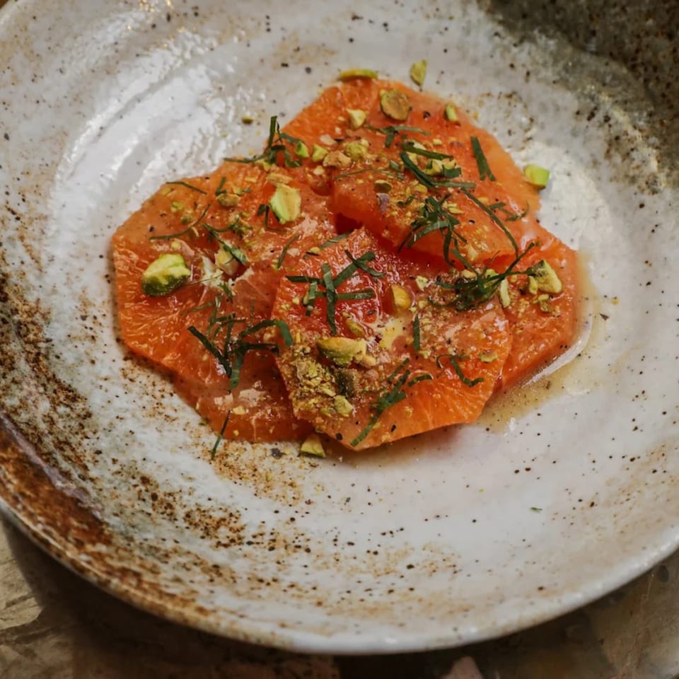 Grapefruit carpaccio with chopped pistachios and fresh herbs on a rustic white plate.