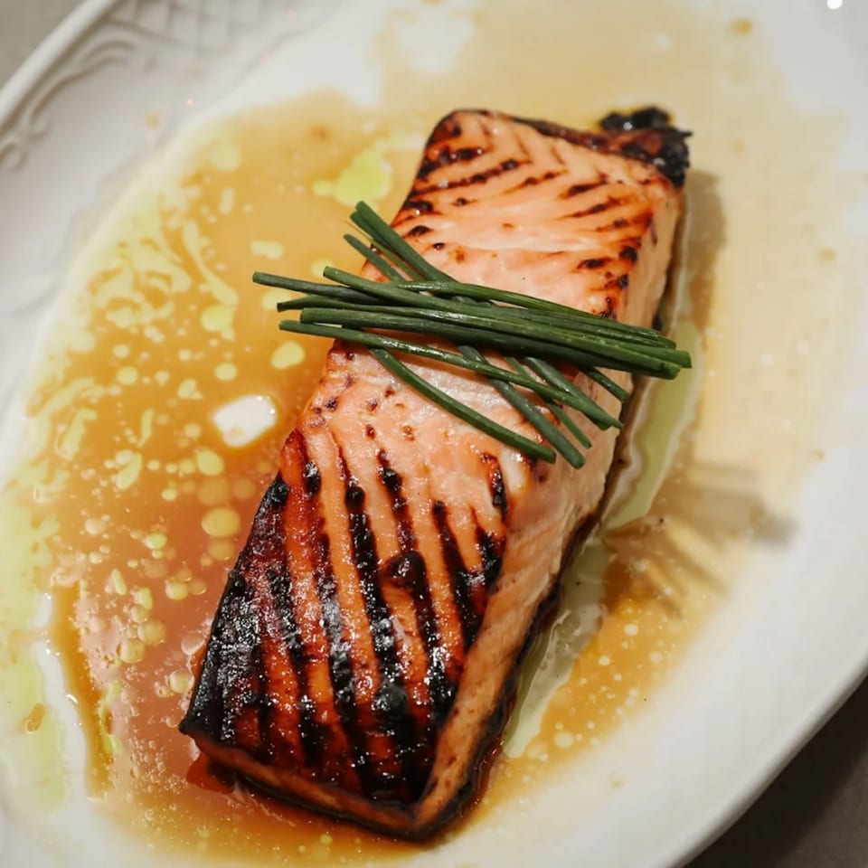 Grilled salmon fillet with grill marks, topped with chives, in a light sauce on a white plate.
