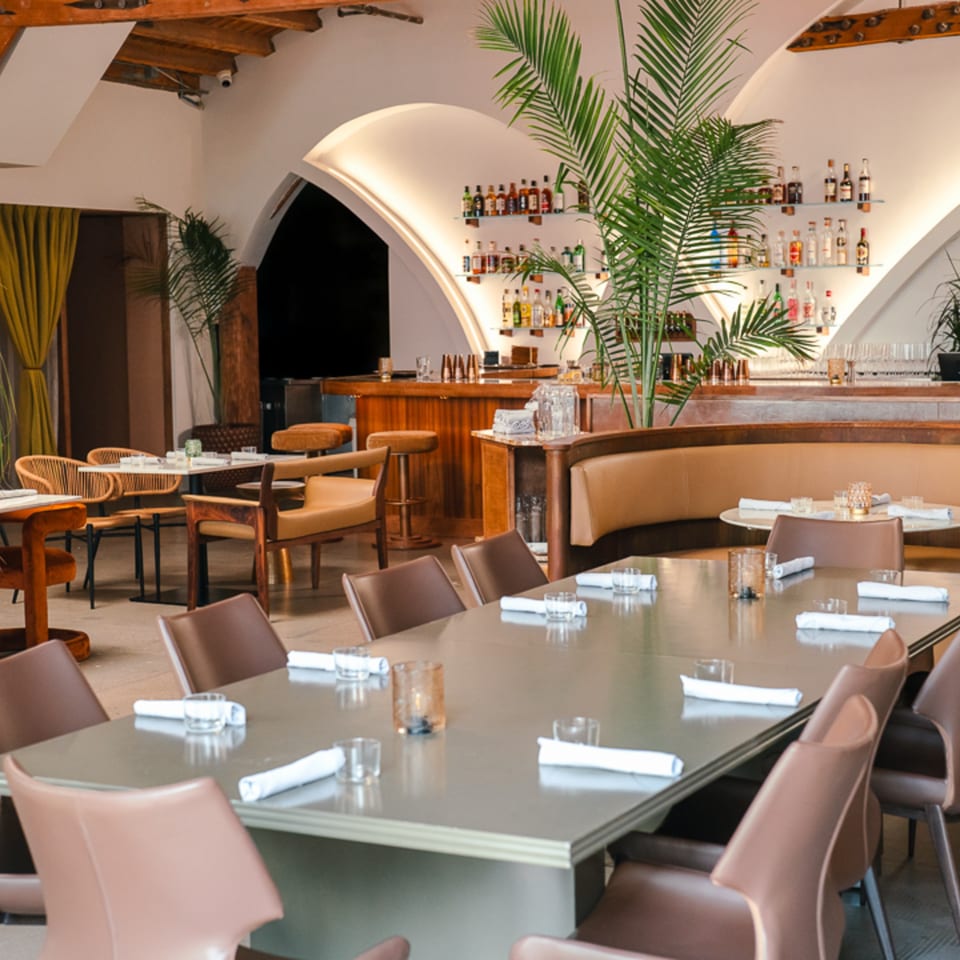 Edgemar Restaurant + Lounge interior with a long dining table, bar, and arched wall shelves.