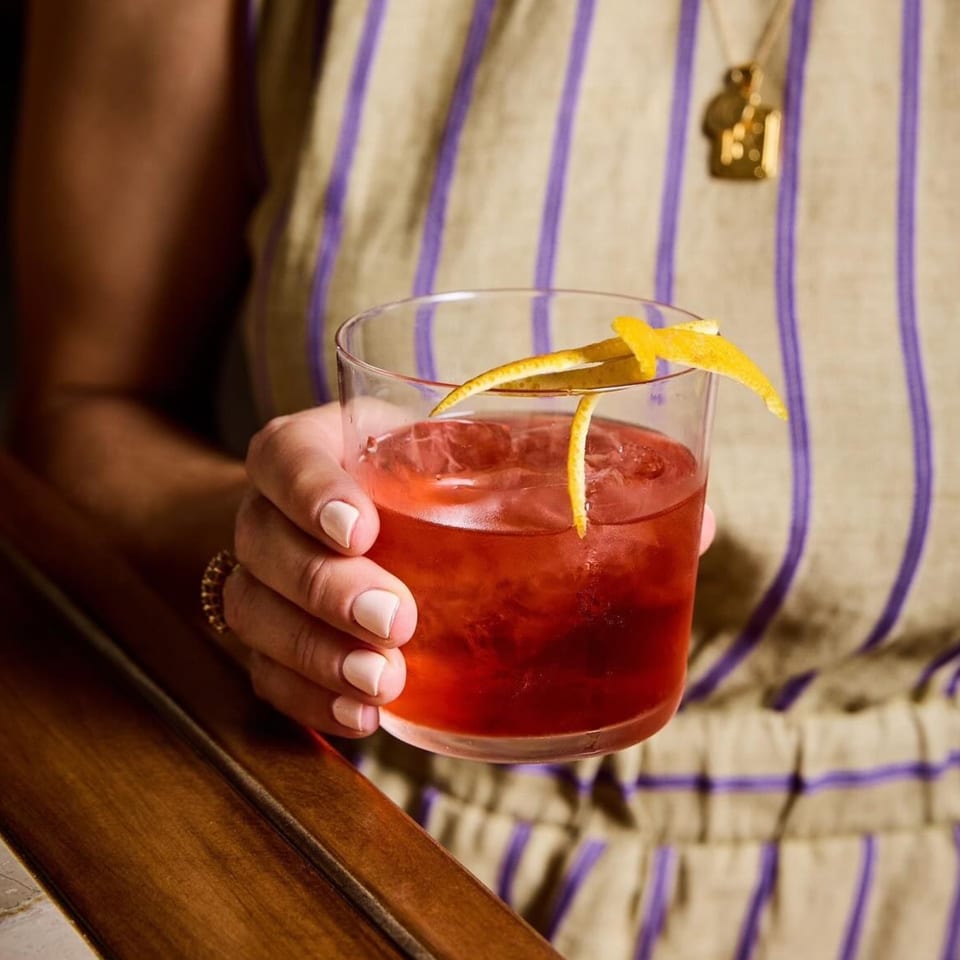 A person in a striped top holds a red cocktail garnished with a lemon peel.