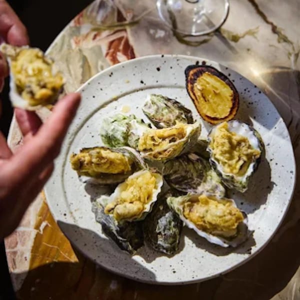 A hand holds an oyster over a plate of baked oysters and a charred lemon half.