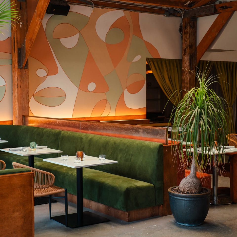 Edgemar Restaurant + Lounge interior with green velvet banquettes, white tables, and a large potted plant.